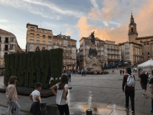 City centre of Vitoria-Gasteiz featuring shrubbery cut into the name of the town, followed by an exclamation point, historic buildings with balconies, and people walking and playing near small fountains.